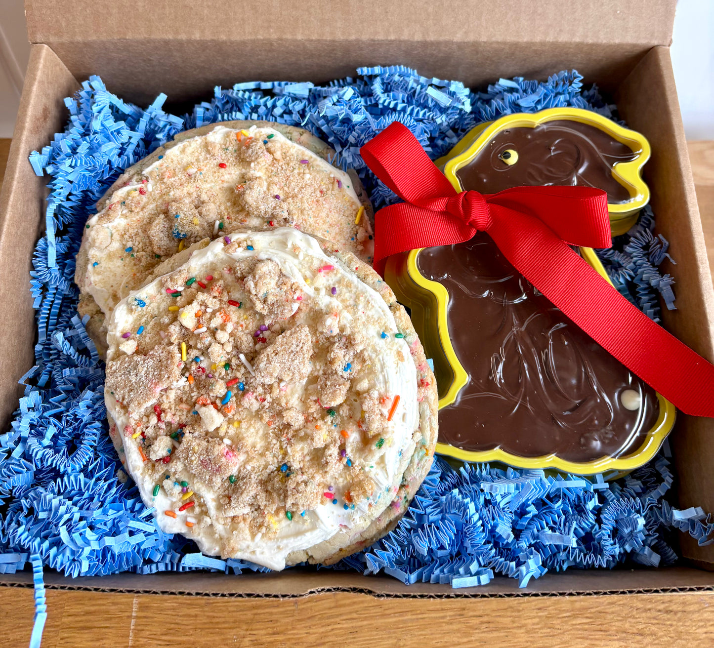 Two frosted sprinkle cookies in a gift box with blue crinkle paper and a chocolate fudge bunny with a red bow.